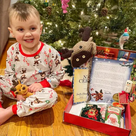 A smiling toddler in reindeer pajamas sitting next to his opened Santa Letter Package under the tree