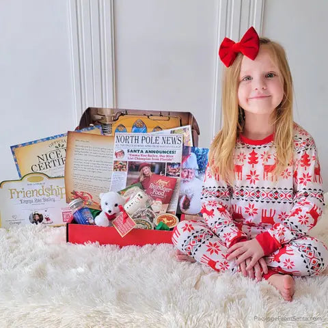 A girl with a red hair bow sitting beside a Santa Letter Package with North Pole News and a Nice List Certificate